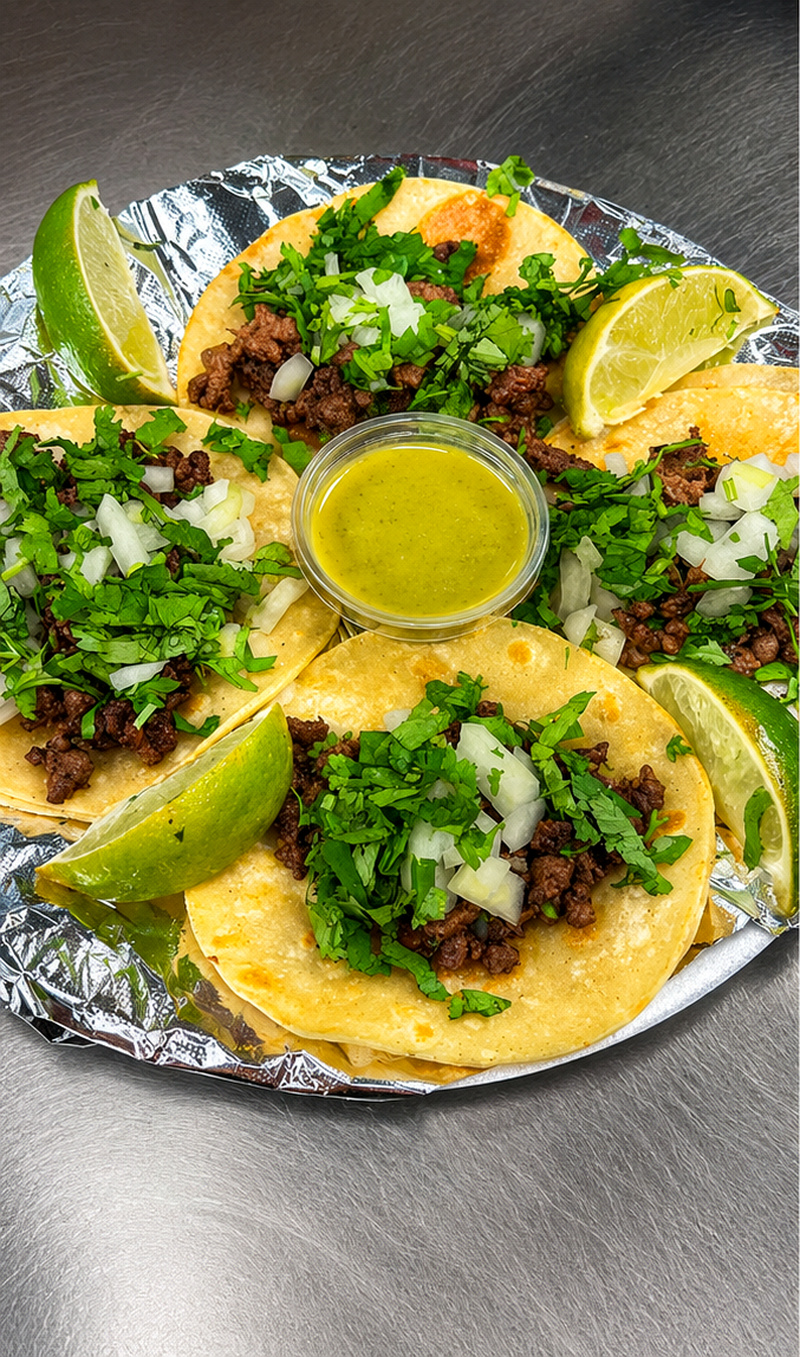 Four street tacos on a platter with green salsa and lime wedges